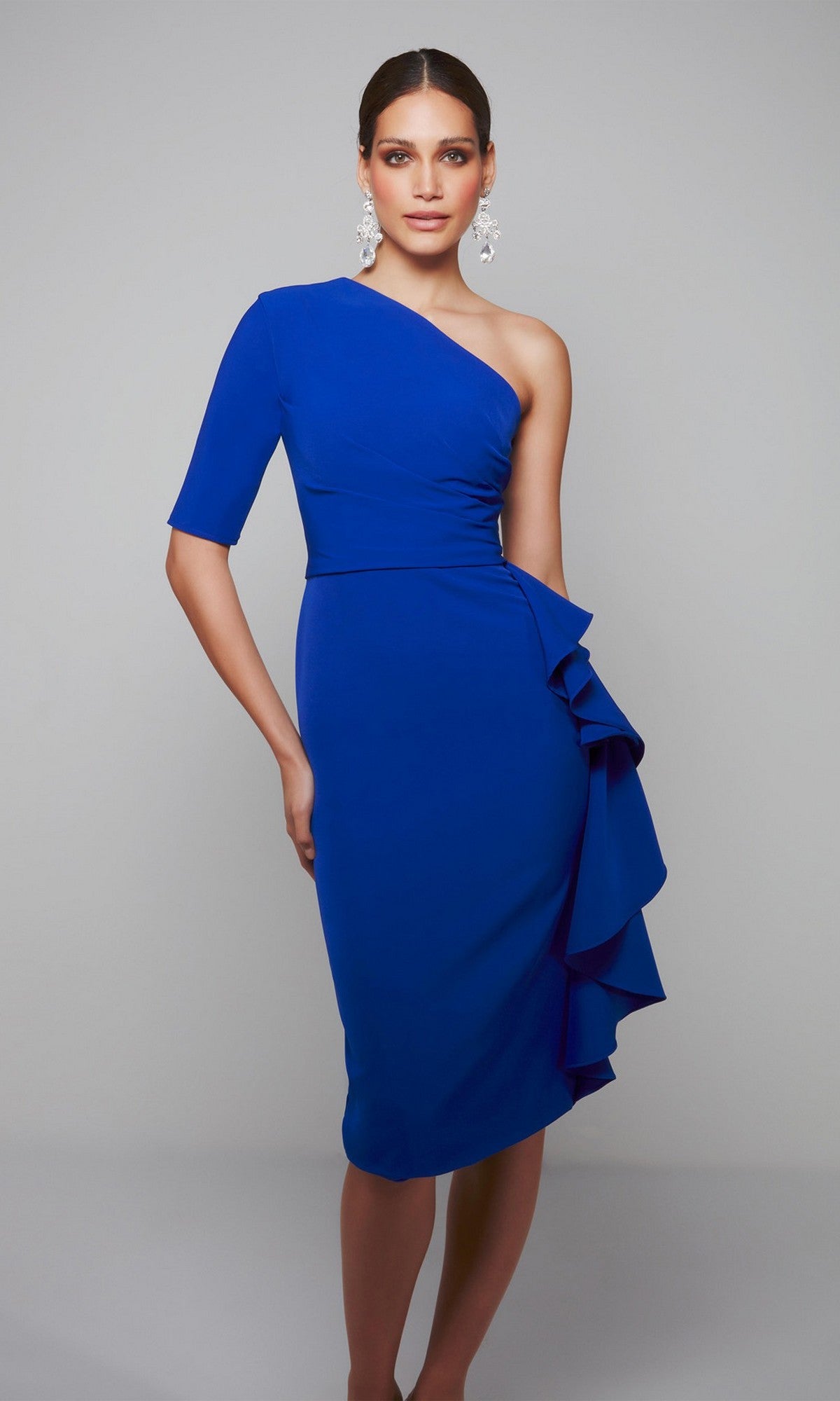 A woman stands against a plain gray background wearing the Alyce 70006 knee-length semi-formal dress, featuring a blue one-shoulder design with ruffle detail on the side.